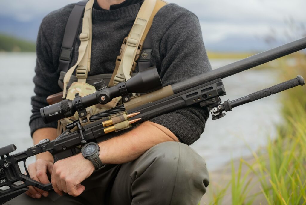 Man sitting in front of a pond with grass holding a large gun